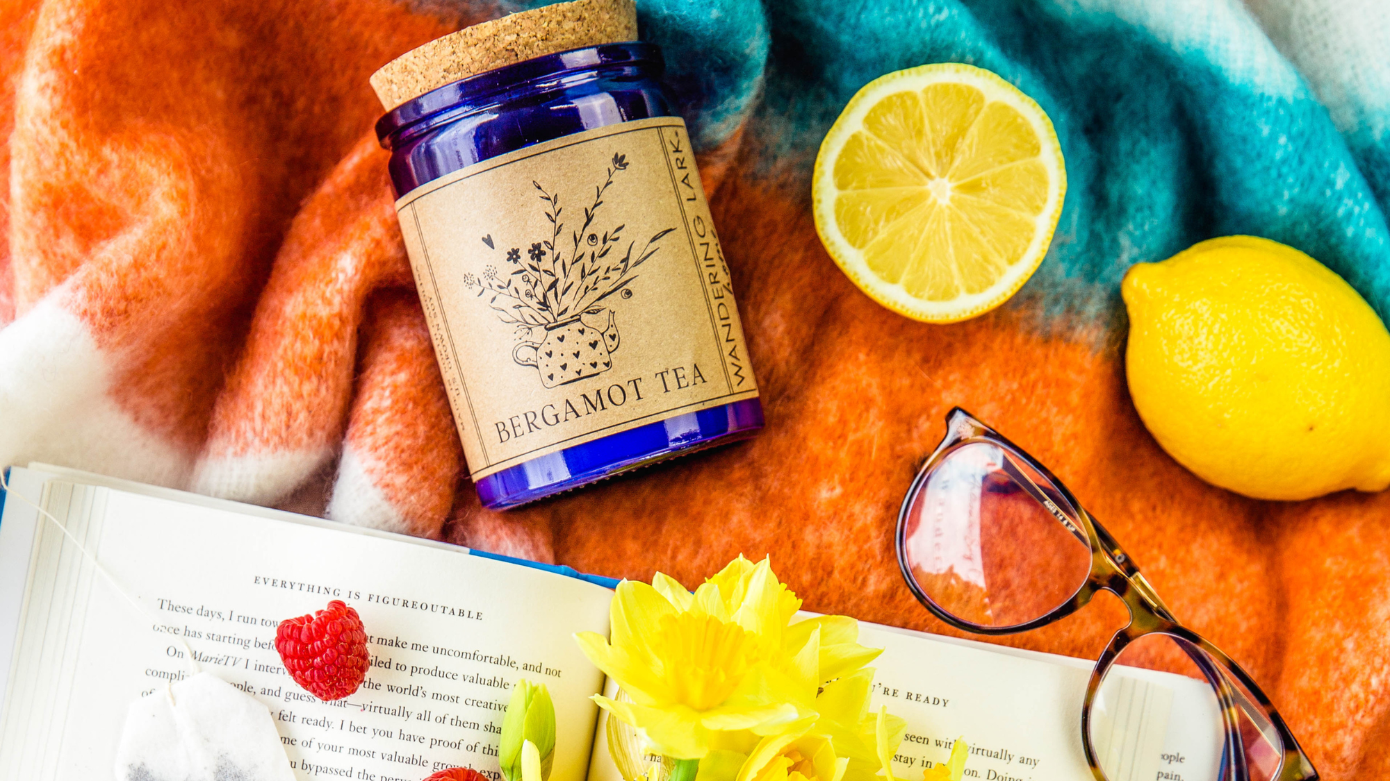 Candle labeled 'Bergamot Tea' with lemons, glasses, and a book on a colorful blanket.