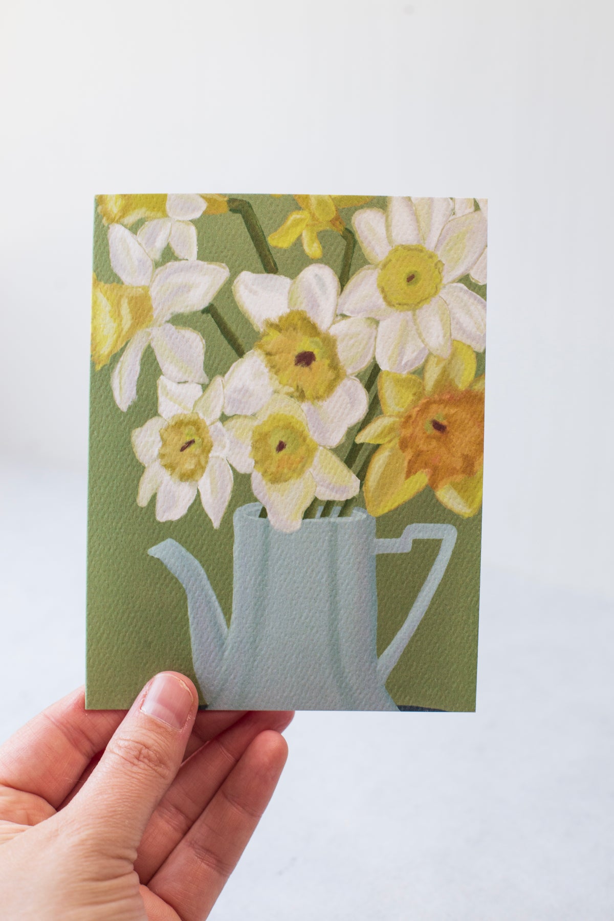 A greeting card on textured paper.  White and Yellow Daffodils are in a blue tea pot in front of a green background.
