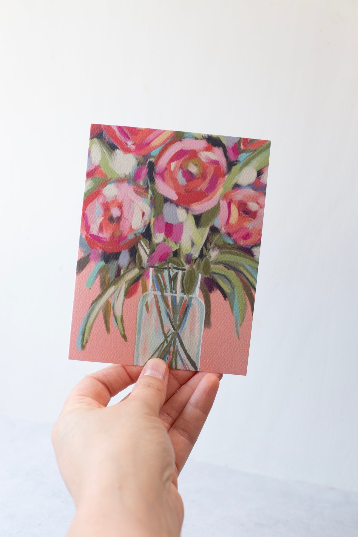 A hand holding a peony-themed greeting card with a pink background and multicolored flowers, printed on textured paper.