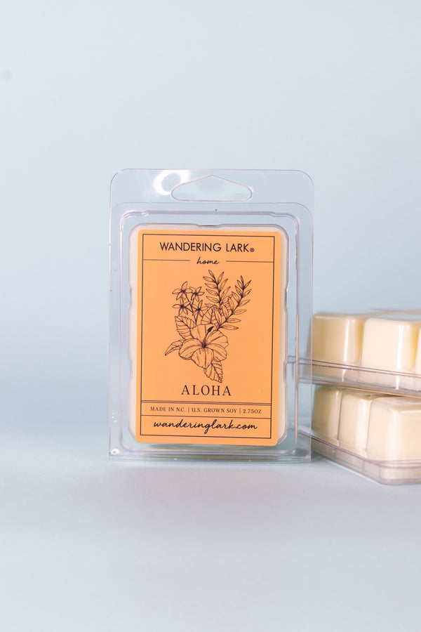 A package of 'Wandering Lark Aloha' wax melts with a few cubes visible in front. The package has a floral and tree illustration and is set against a light blue background.