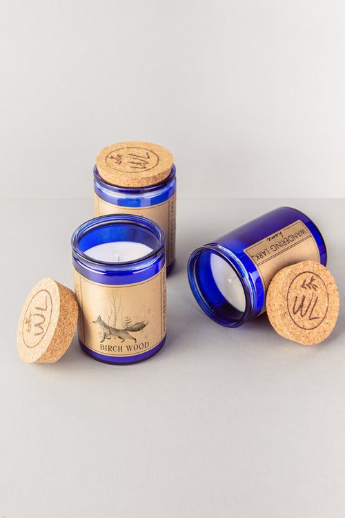 Three soy candles with cork lids, one with the lid removed, revealing a candle inside. The other two have labels with 'Birch Wood' written on them and an illustration of a fox in a wood.  The jar is blue