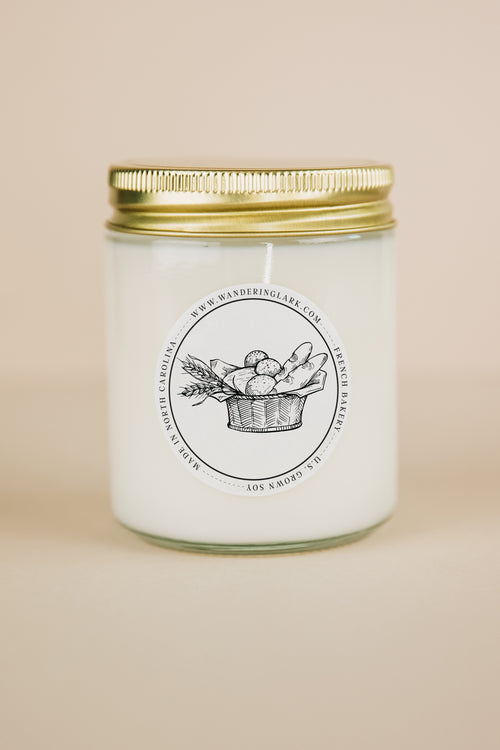 French Bakery scented candle in a clear jar with a gold lid.  The label is white and round with an illustration of bread in a basket.