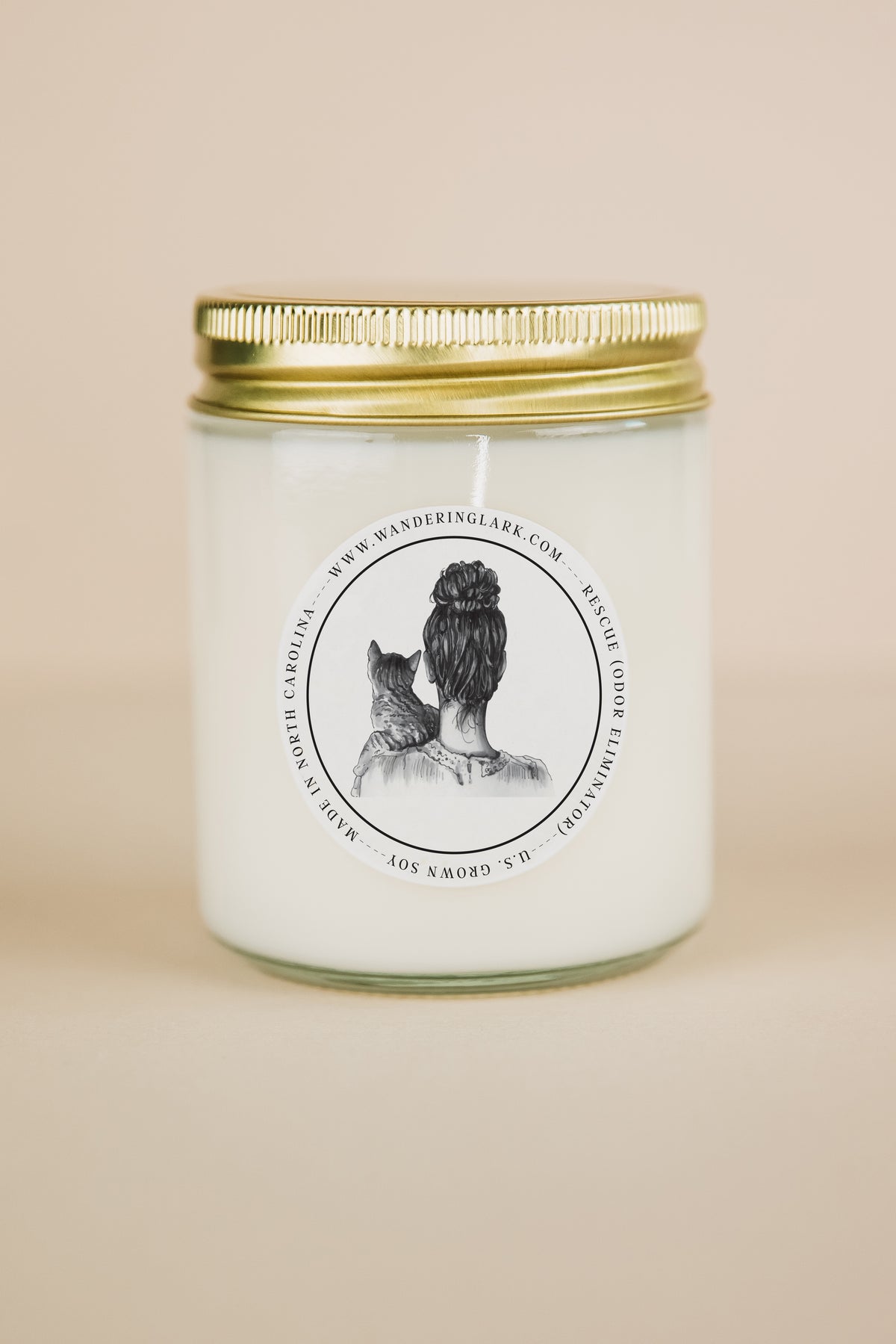 A clear glass candle, gold lid, white wax, with a label that has a graphic of a person holding a cat, with text 'Rescue (odor eliminator)'. 