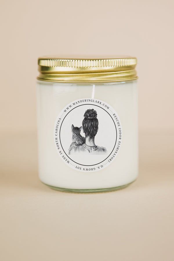 A clear glass candle, gold lid, white wax, with a label that has a graphic of a person holding a cat, with text 'Rescue (odor eliminator)'. 