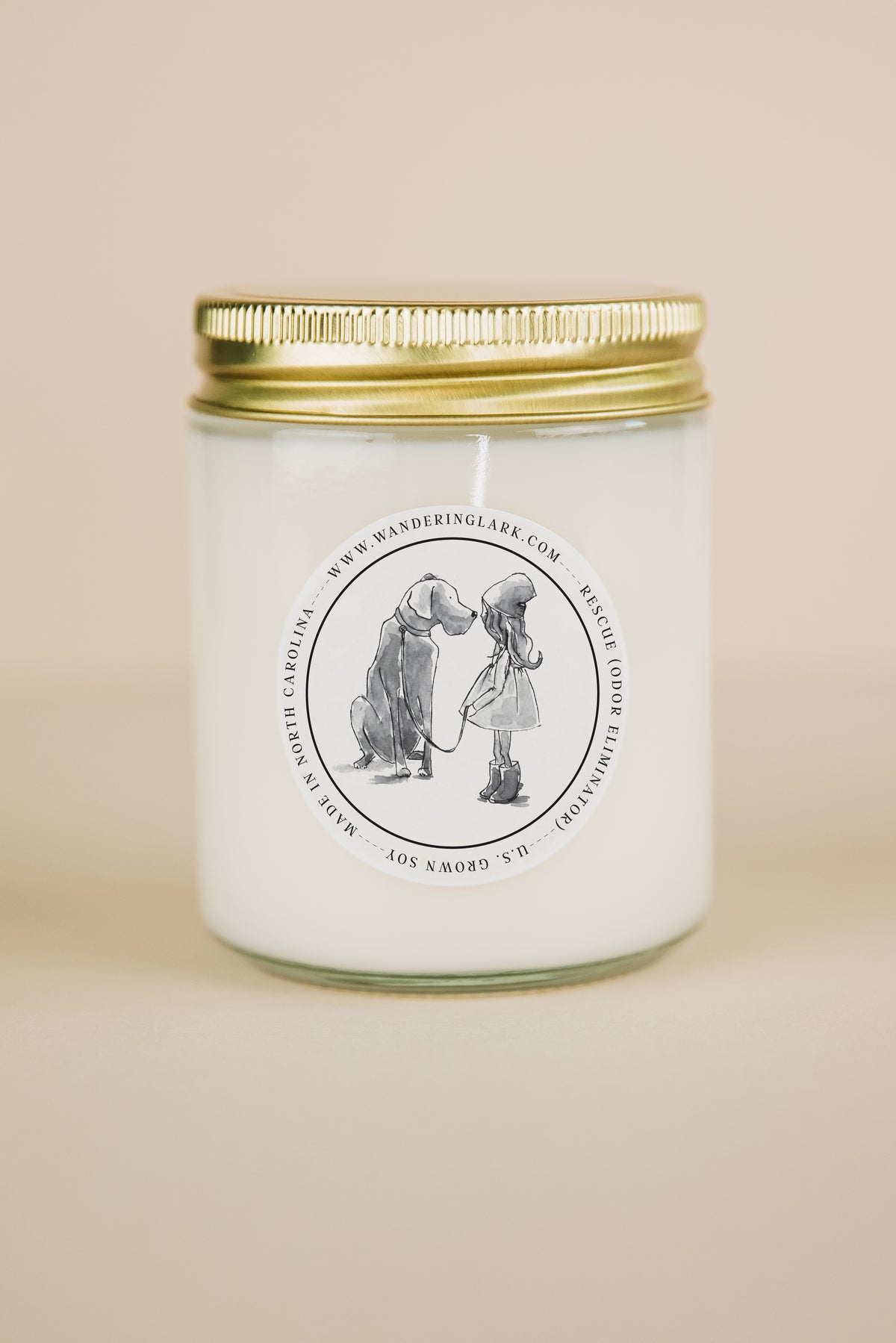 A clear glass candle, gold lid, white wax, with a label that has a graphic of a person crouching down and a dog sitting up, with text 'Rescue (odor eliminator)'. 