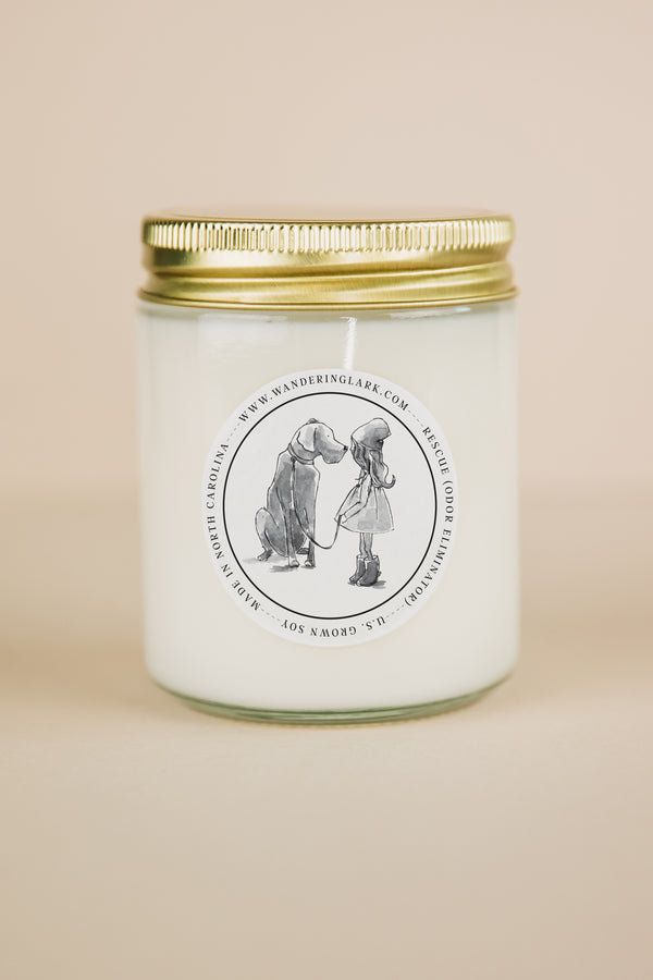 A clear glass candle, gold lid, white wax, with a label that has a graphic of a person crouching down and a dog sitting up, with text 'Rescue (odor eliminator)'. 