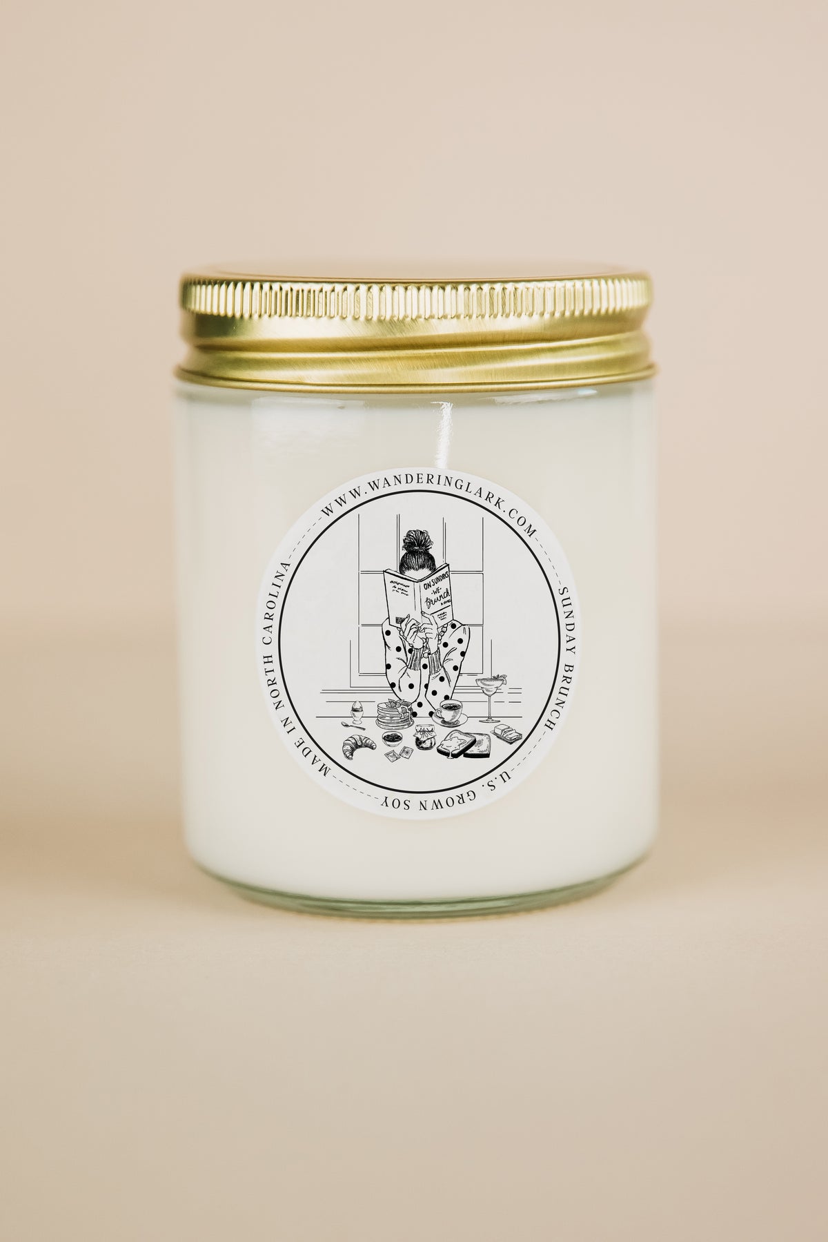A clear glass candle, gold lid, white wax, with a label that has a graphic of gril at brunch reading a book.