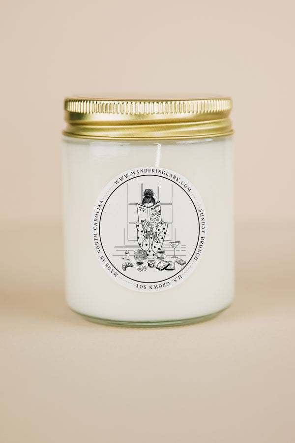 A clear glass candle, gold lid, white wax, with a label that has a graphic of gril at brunch reading a book.