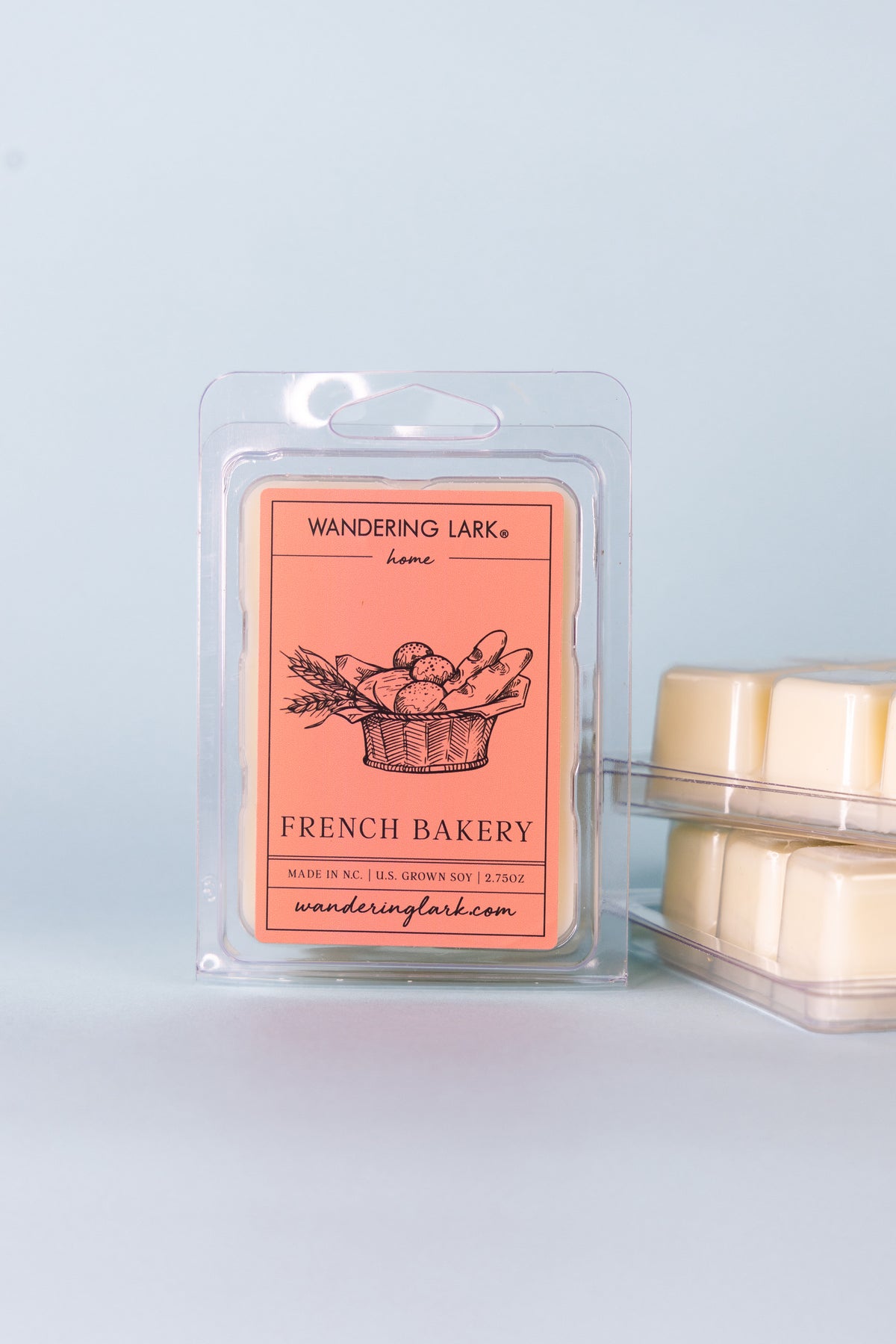 A package of Wandering Lark French Bakery wax melts, with a clear window showing the orange label and the scent profile of clove, cinnamon, and vanilla.