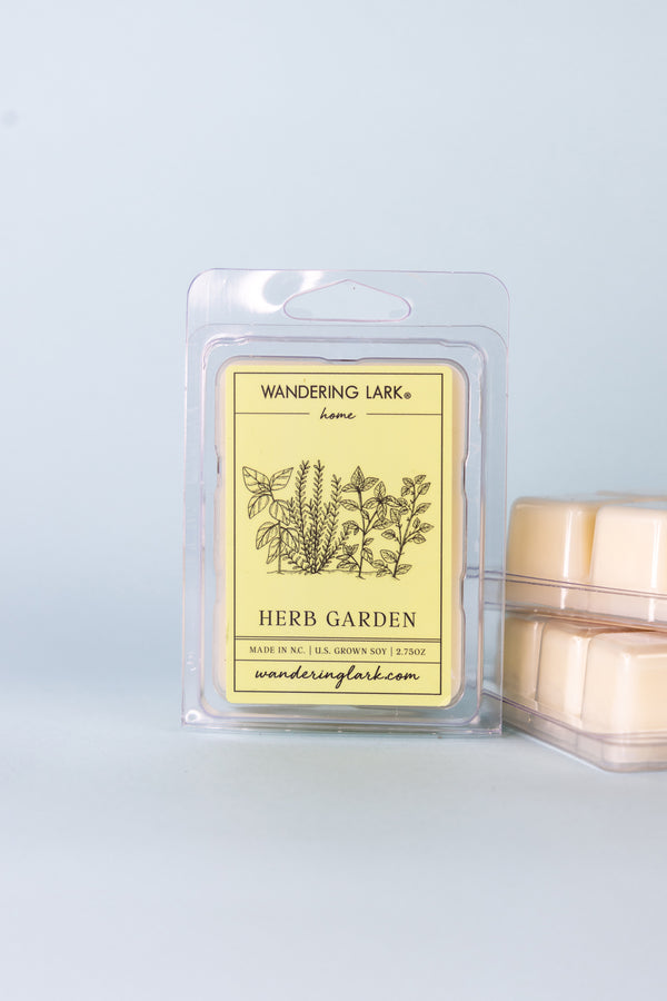 A package of 'Herb Garden' wax melts by Wandering Lark, featuring a clear plastic container with a yellow label that has botanical illustrations and the brand's website.