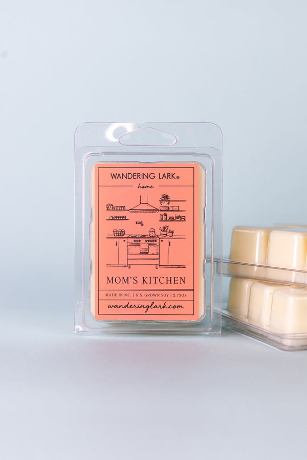 A package of 'Mom's Kitchen' wax melts by Wandering Lark, with the product visible through a clear plastic front. The packaging is red with an illustration of a kitchen stove and shelves, and there are wax melt cubes showing on the back.