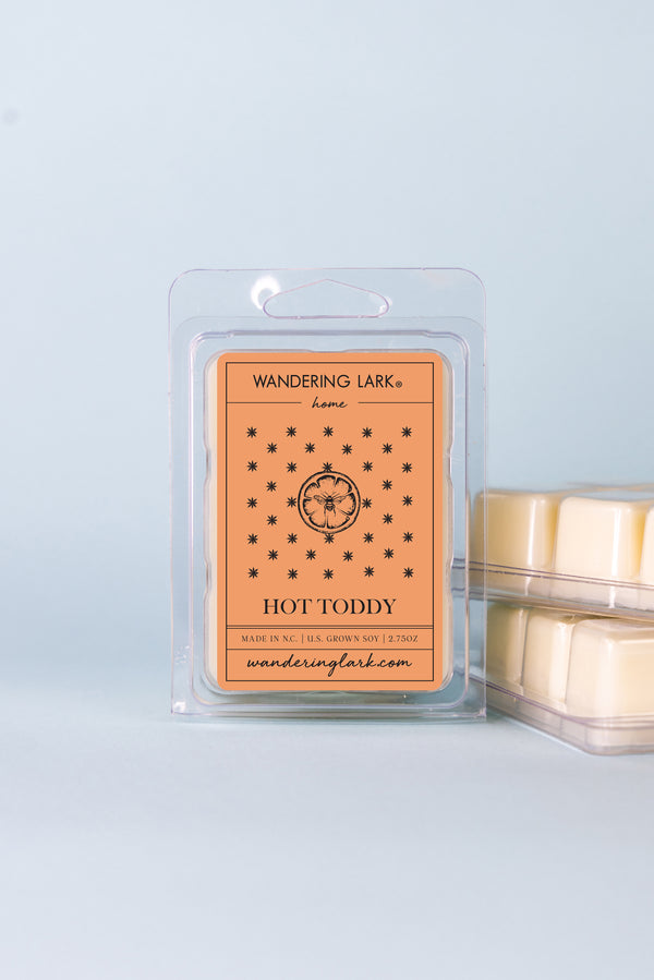 A package of Hot Toddy wax melts by Wandering Lark, with visible wax cubes in a clear container against a blue background.